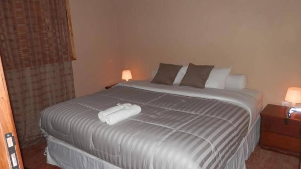 Una cama bien hecha con edredón gris, dos almohadas y una toalla enrollada en el cabecero. Hay dos mesitas de noche con lámparas.