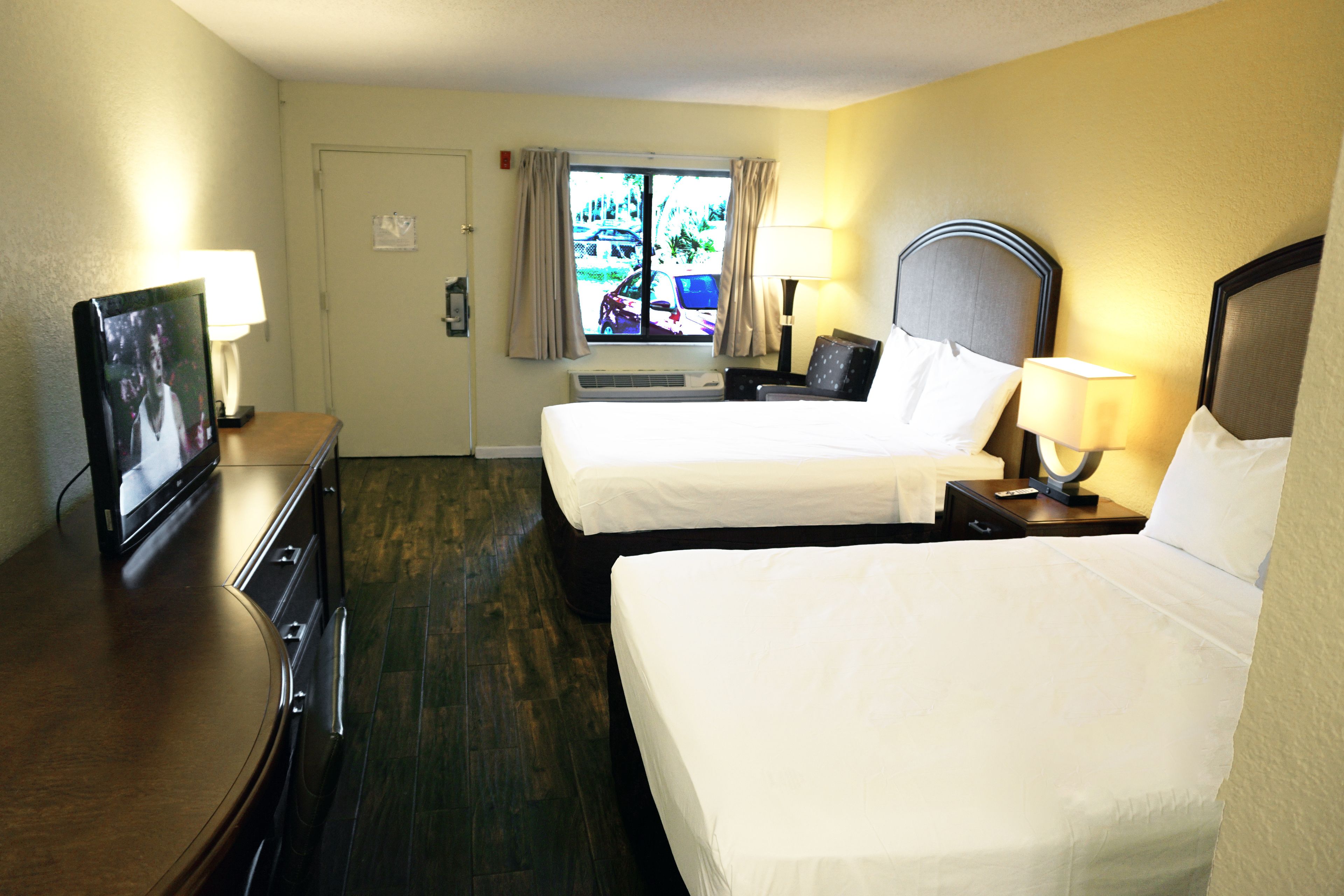Habitación de hotel con dos camas, un escritorio con televisor, una ventana con cortinas y una puerta.