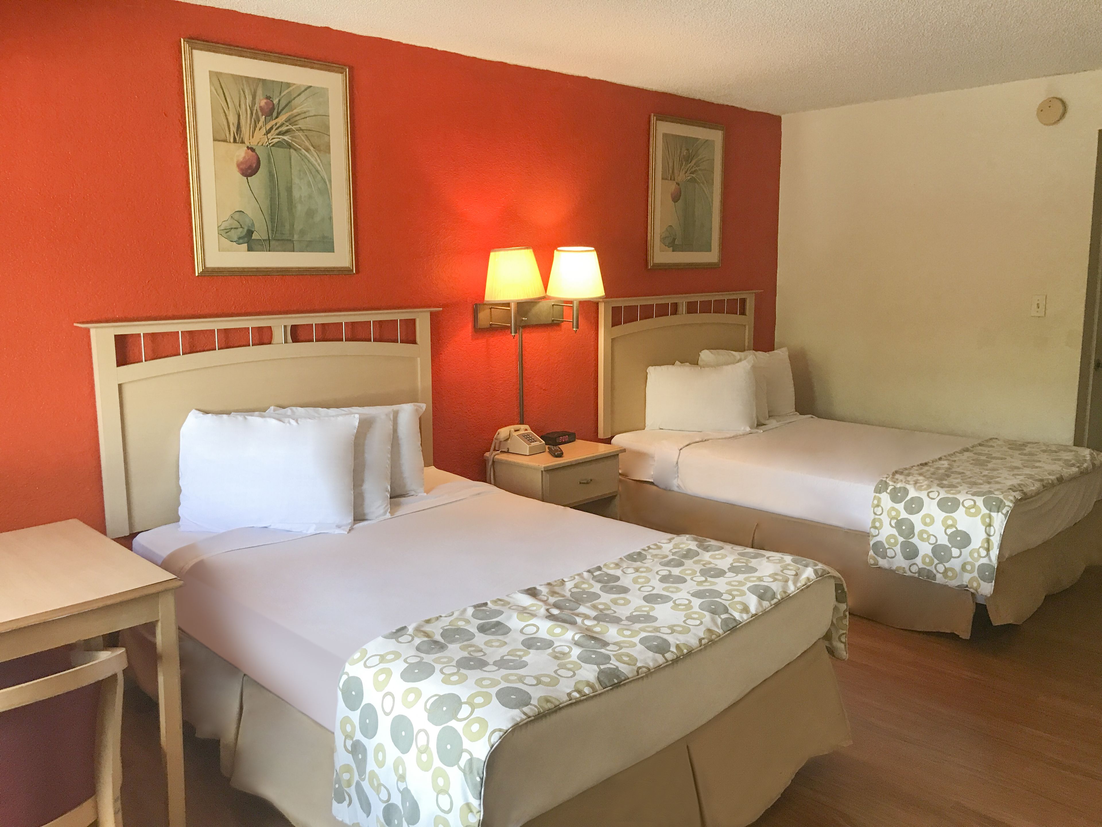 A hotel room with two beds, a nightstand with a lamp, and two framed floral paintings on the wall.