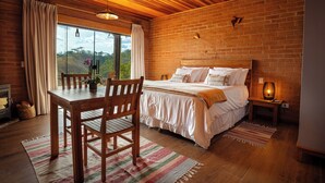 A bedroom with a wooden ceiling, brick walls, a large bed, a wooden table with chairs, and a view of the outdoors through a window. - VELINN Hotel Fazenda Fonte das Hortênsias (Santo Antonio do Pinhal)