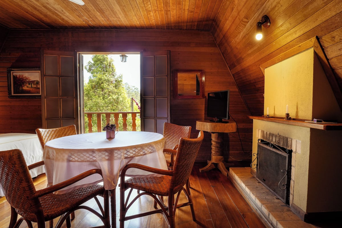 A cozy room with a wooden interior, a round table with chairs, a fireplace, and a large window with shutters.