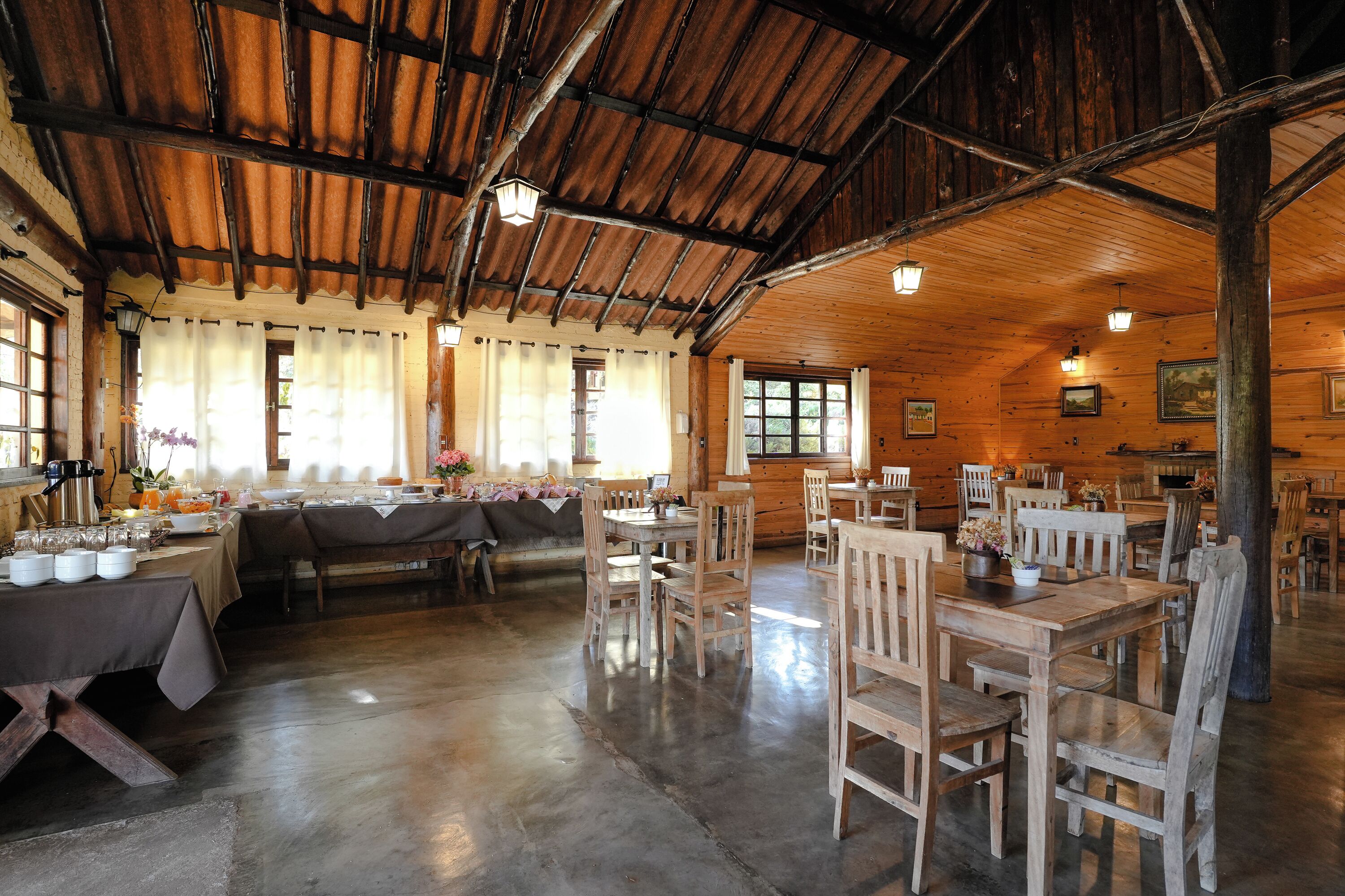 Un'accogliente sala da pranzo in stile rustico, con tavoli e sedie in legno, ampie finestre con tende e un alto soffitto in legno.