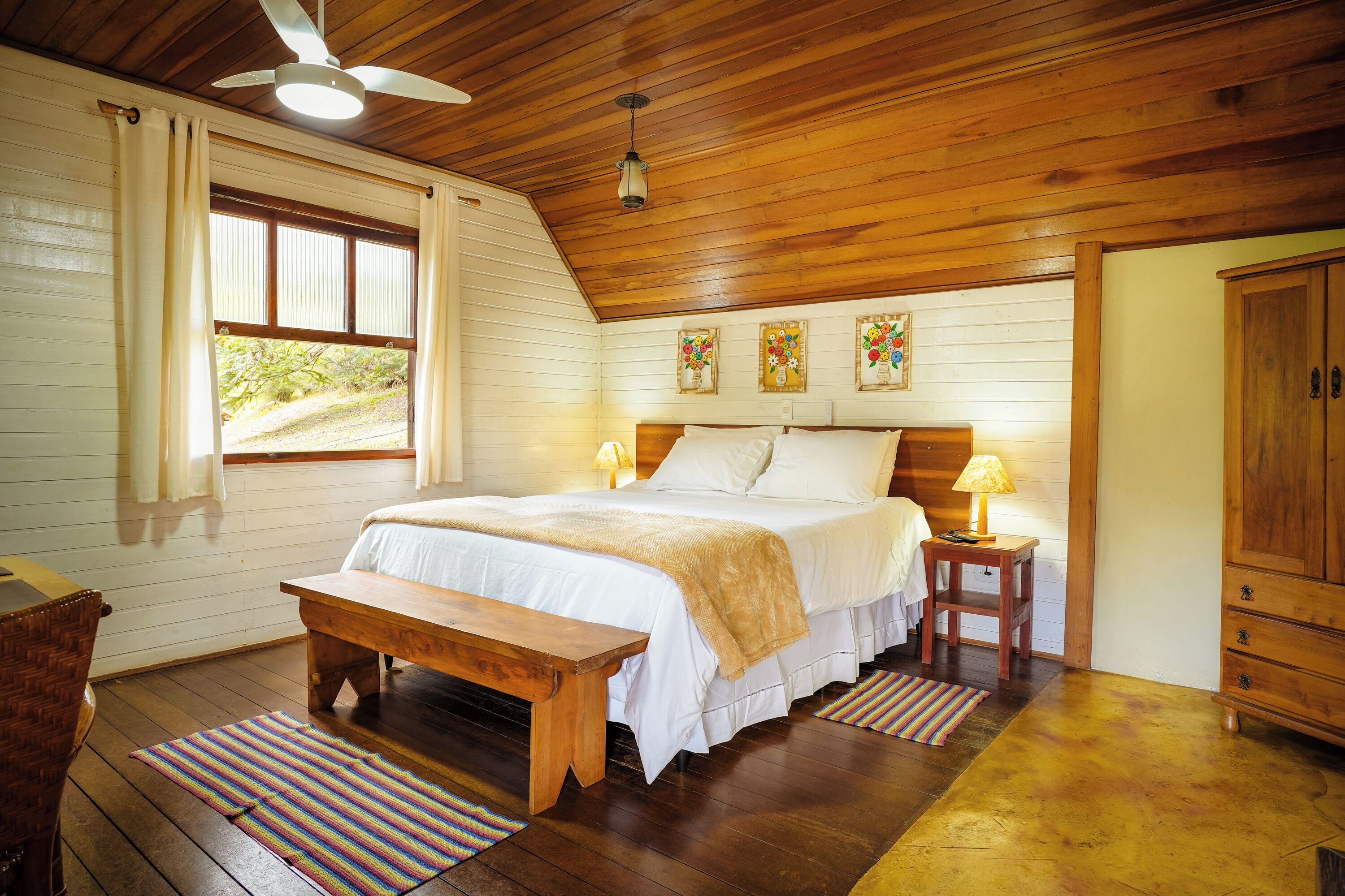 A bedroom with a wooden ceiling, a bed with white bedding, a bench, a bedside table with a lamp, and a window with curtains.