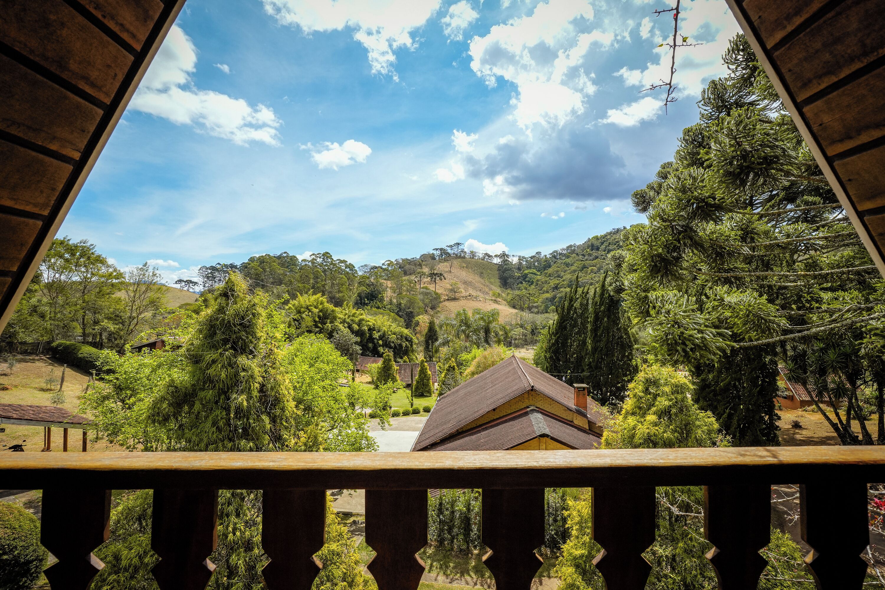 A view from a balcony with a wooden railing, overlooking a green landscape with trees and a hill in the distance.