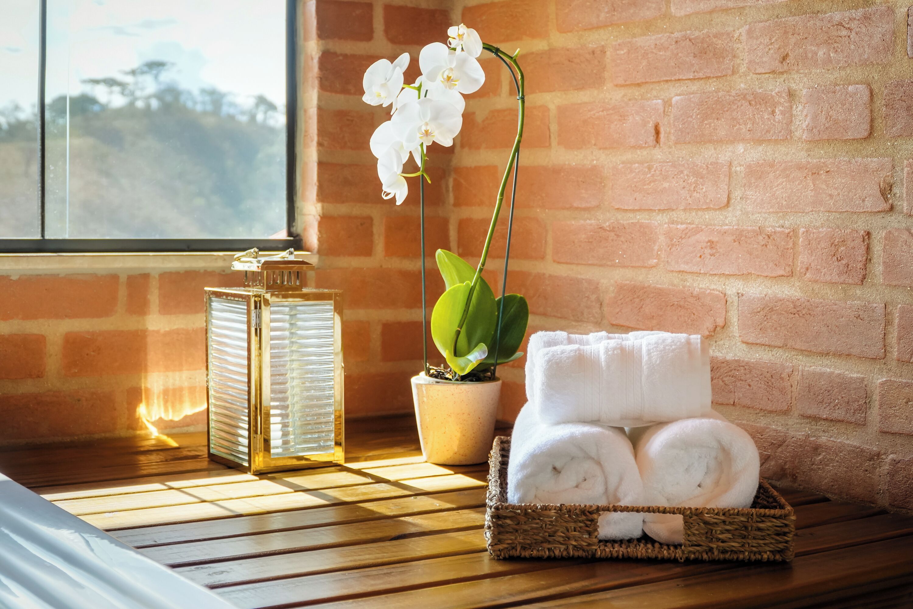 A wooden bench with a potted orchid, a glass bottle, and a basket of rolled towels against a brick wall.
