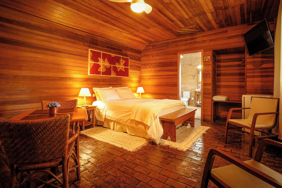 A wooden-floored bedroom with a bed, two bedside lamps, a flat-screen TV mounted on the wall, and a dining area with a table and chairs.