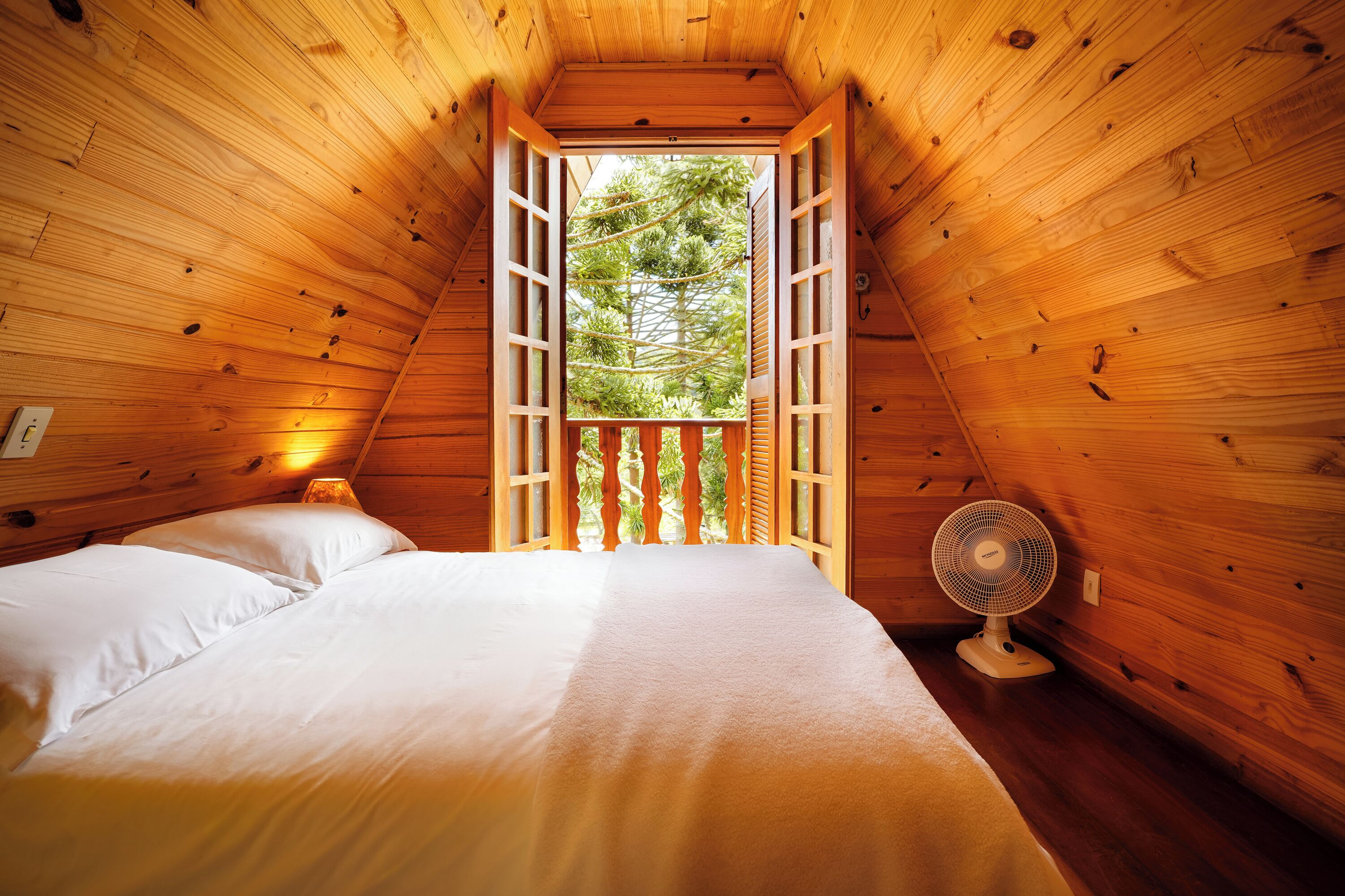 Acogedora habitación tipo cabaña de madera con cama, ventilador y ventana con vistas a árboles.