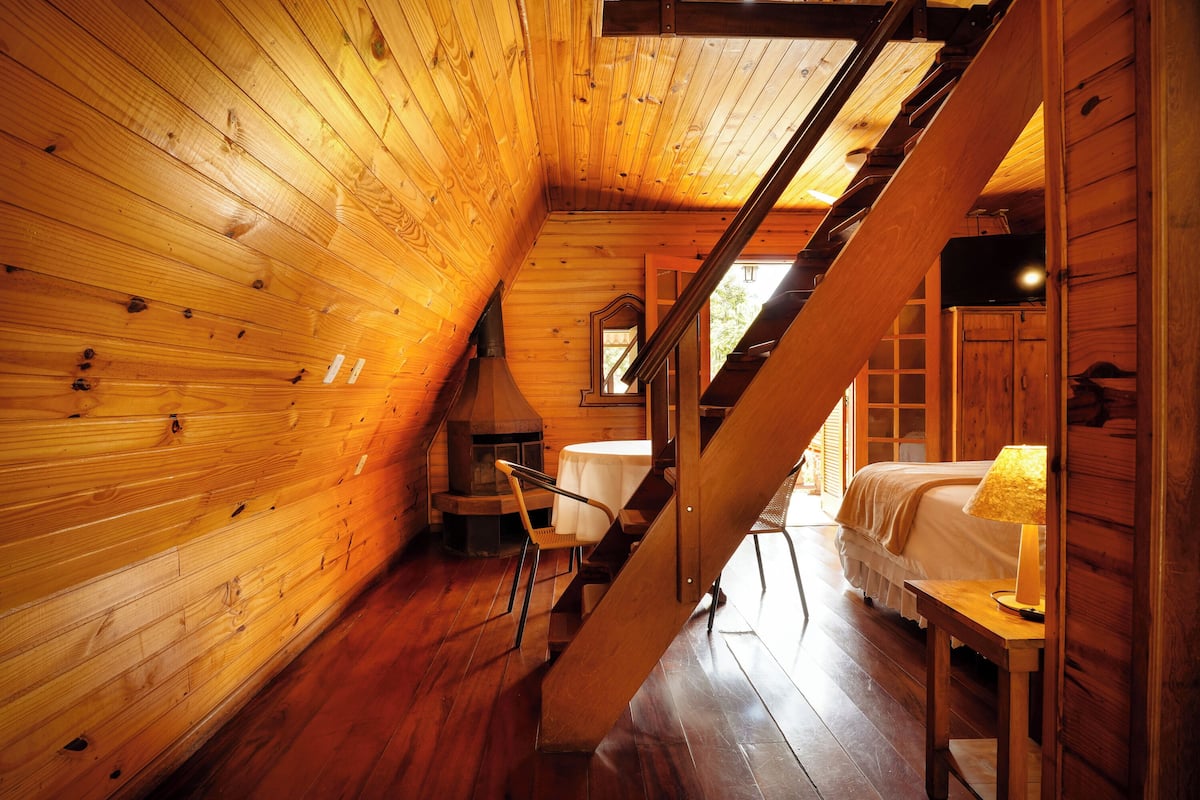 A wooden cabin interior with a staircase, a bed, a table, and a chair.