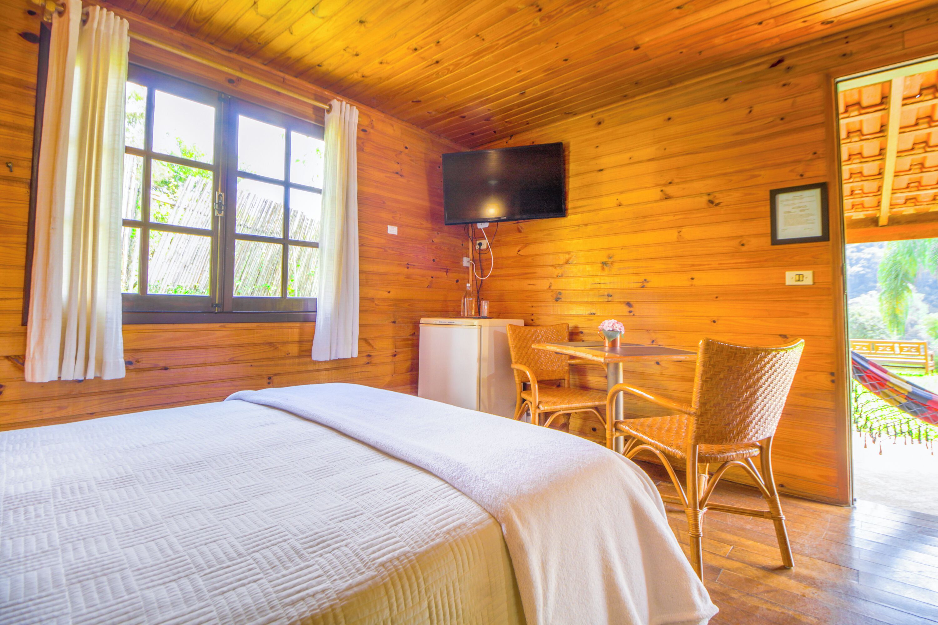 A wooden room with a bed, a TV mounted on the wall, a small table with chairs, and a window with curtains.