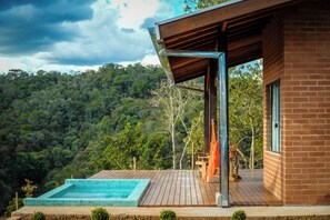 Une maison moderne dotée d’une terrasse en bois, d’une piscine et offrant une vue sur une colline boisée.
