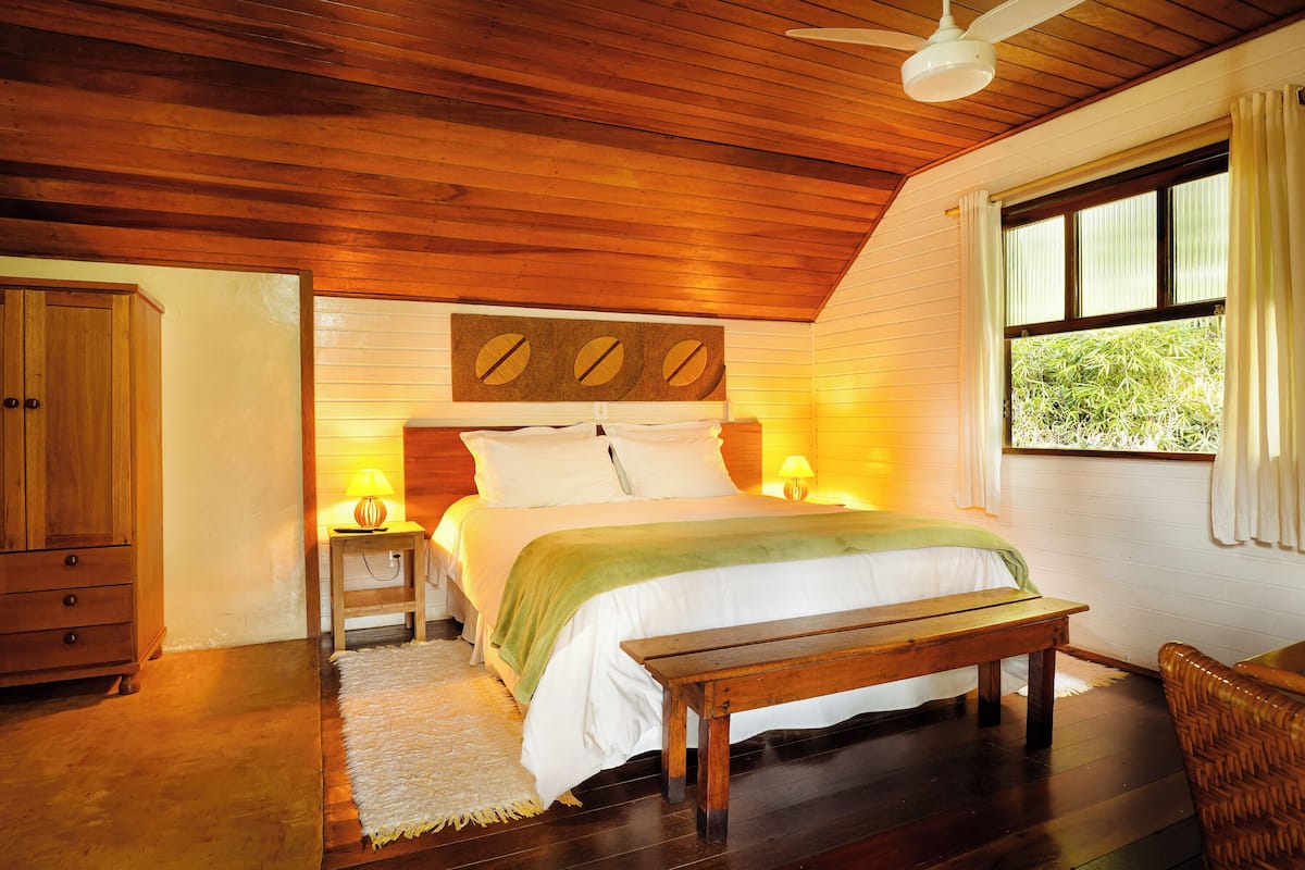 A bedroom with a wooden ceiling, a bed with white bedding, a wooden bench, a nightstand with a lamp, and a window with curtains.