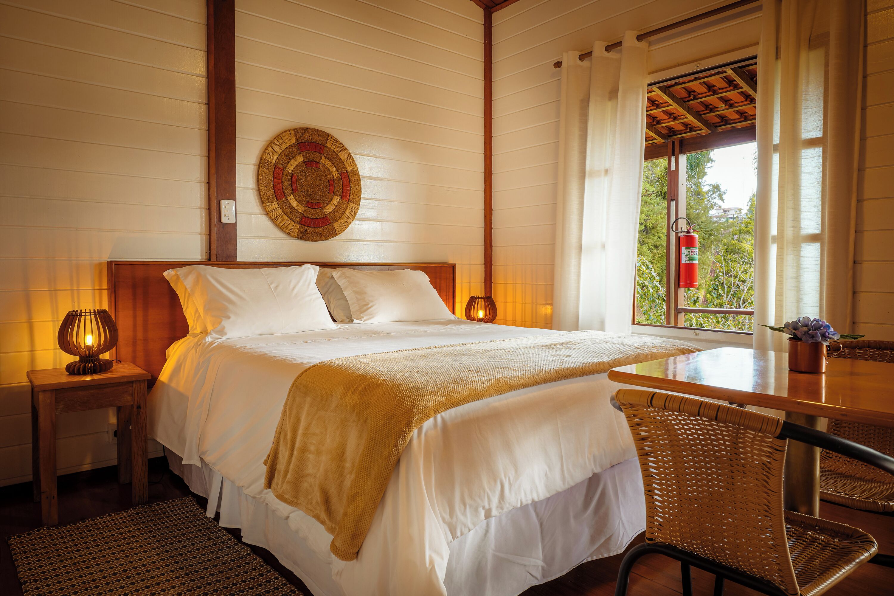 A neatly made bed with white linens, a wooden headboard, and a bedside lamp. There is a wicker chair and a small table with a vase of flowers.