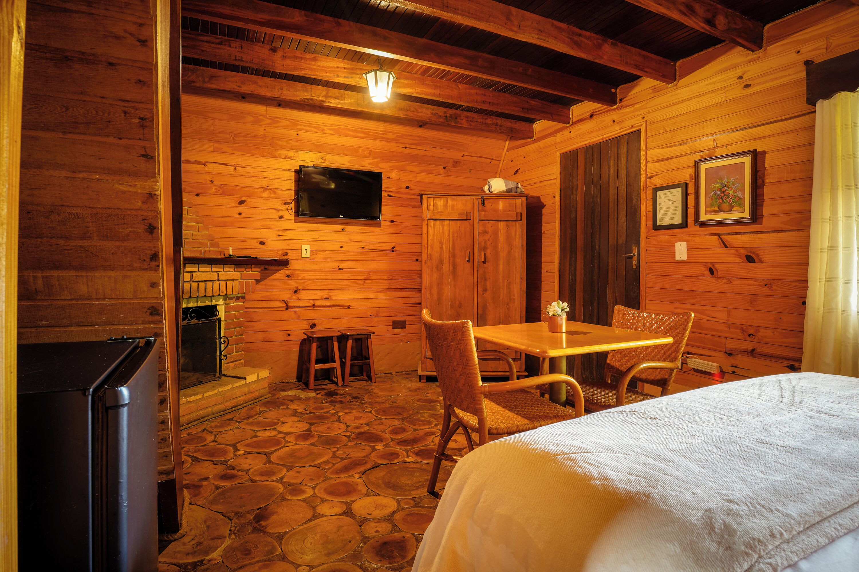 Acogedora habitación tipo cabaña de madera con suelo de piedra, paredes de madera, una cama, una mesa, sillas, un televisor y una chimenea.