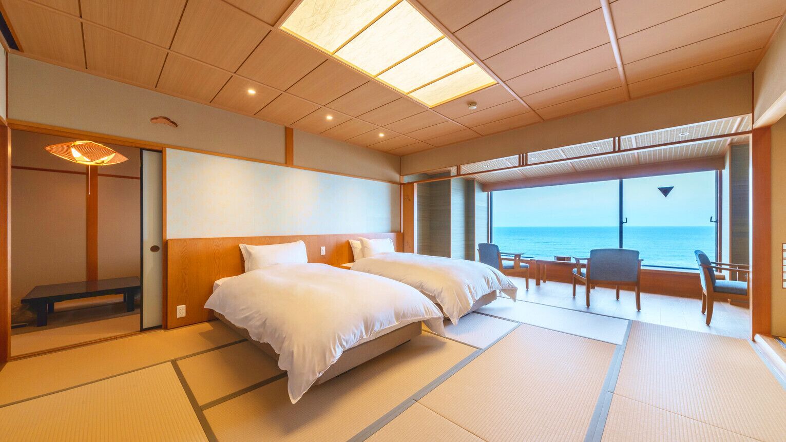A hotel room with a bed, a bench, a table, and chairs near a large window with a view of the sea.