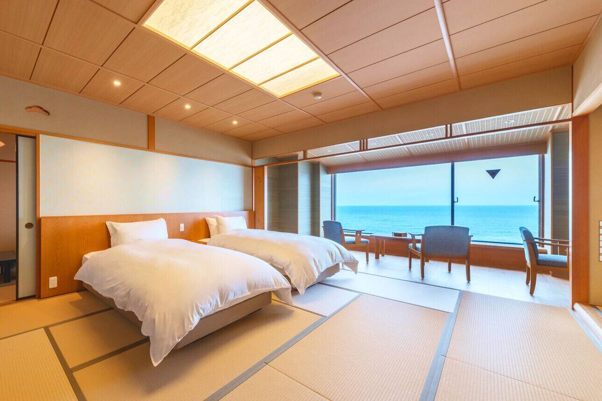 A hotel room with a bed, a bench, a table, and chairs near a large window with a view of the sea.