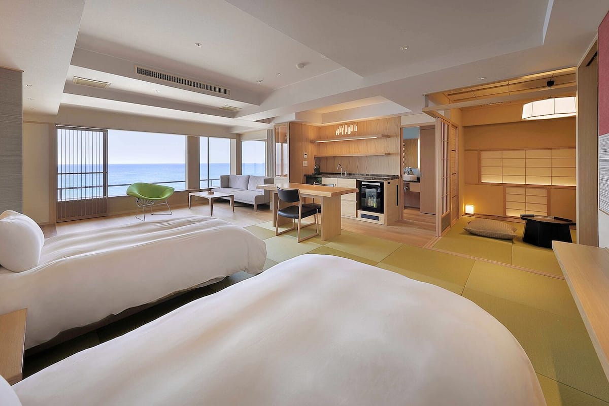 A hotel room with two beds, a kitchenette, a seating area with a view of the sea, and a dining area.