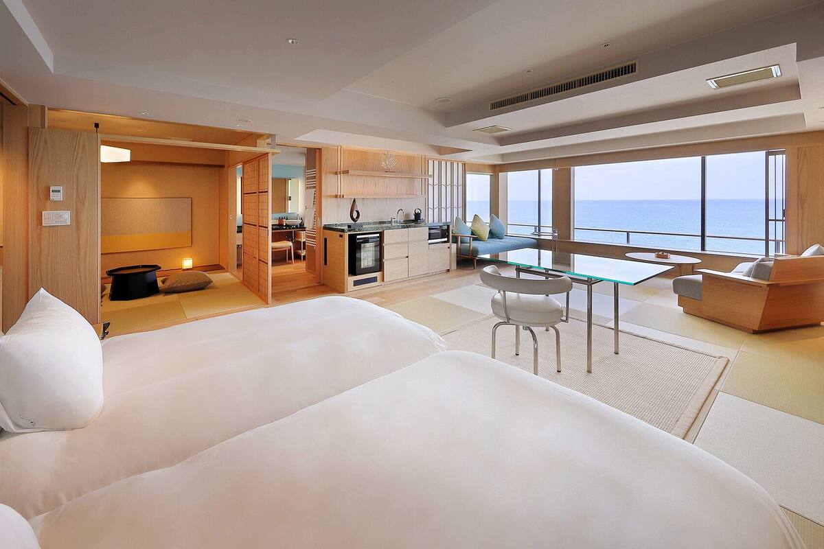 A modern hotel room with a large bed, a dining area, and a view of the sea.