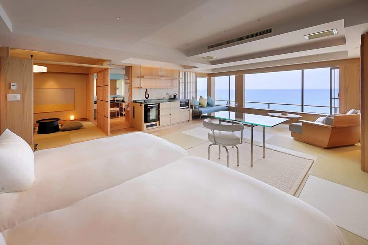 A modern hotel room with a large bed, a dining area, and a view of the sea.
