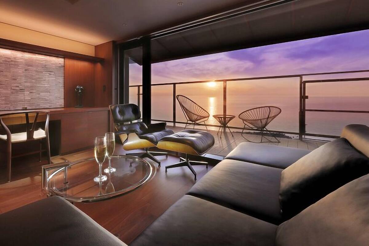 A modern interior with a glass table, leather seating, and a view of the sunset over the ocean.