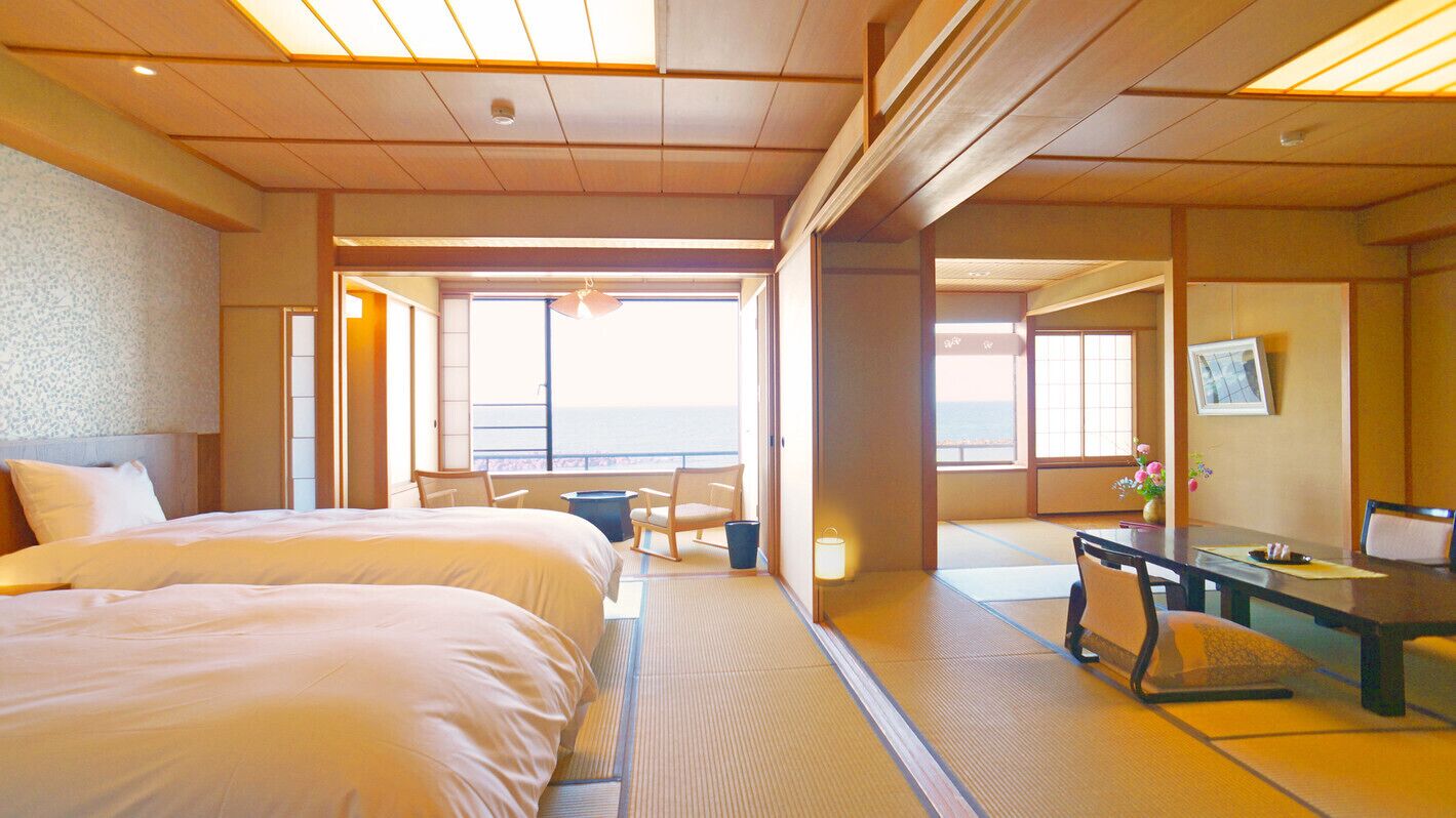 A hotel room with two beds, a desk, a chair, and a view of the sea.
