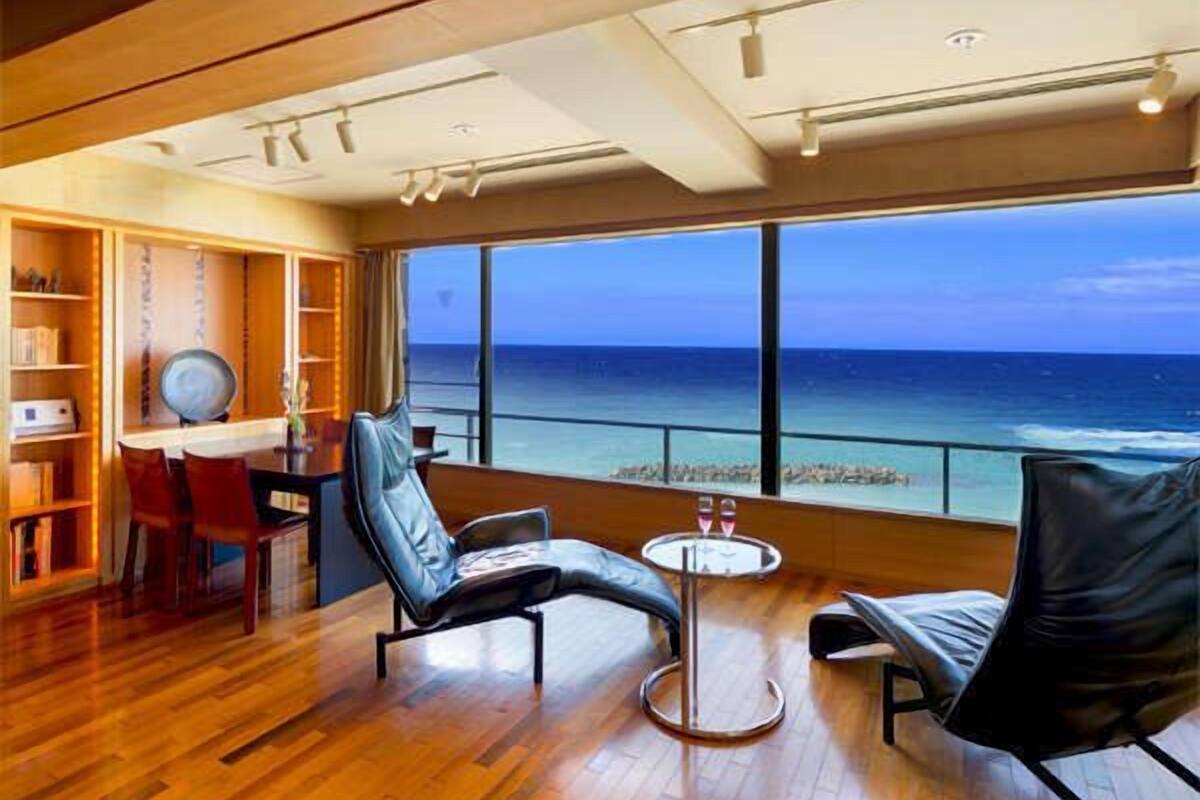 A room with a large window overlooking the ocean, wooden flooring, a seating area with chairs, a small table, and a bookshelf.