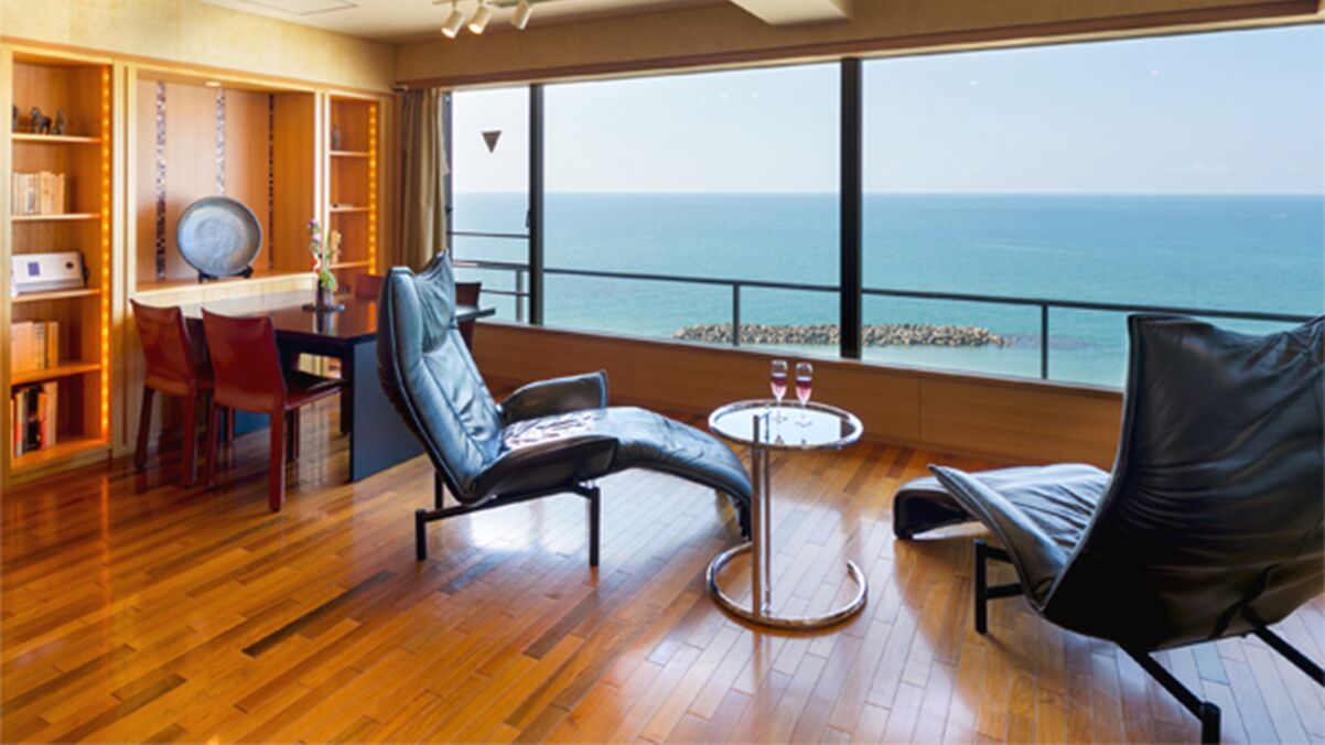 A room with a wooden floor, a dining table with chairs, a sofa, a small round table with two glasses, and a view of the sea through large windows.