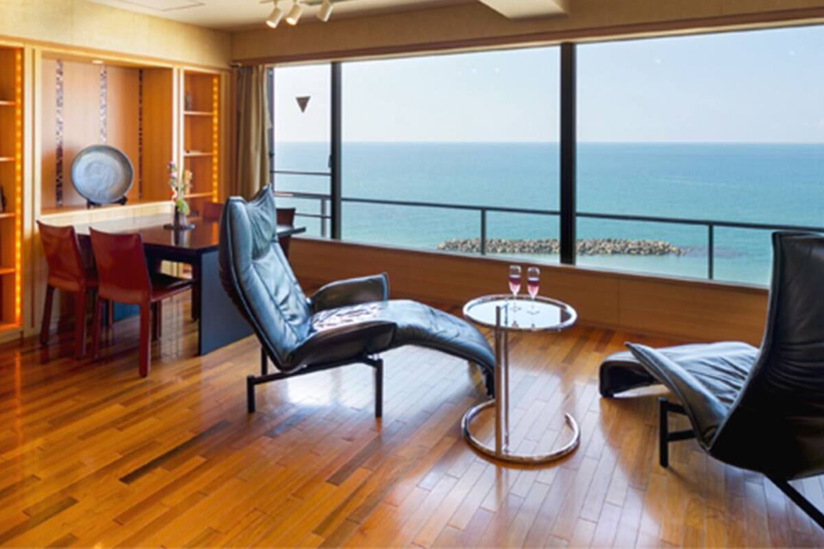 A room with a wooden floor, a dining table with chairs, a sofa, a small round table with two glasses, and a view of the sea through large windows.