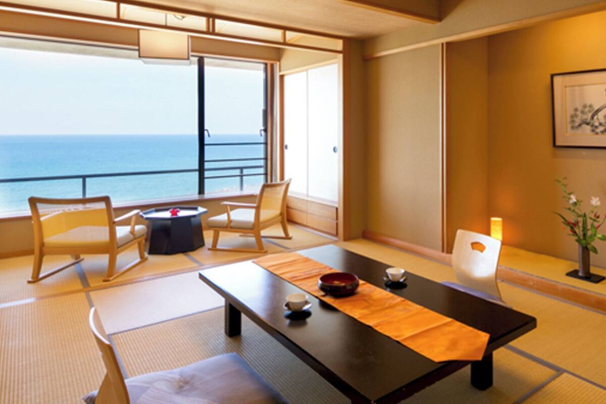 A traditional Japanese room with tatami flooring, a low table, chairs, a view of the sea, and a framed artwork on the wall.