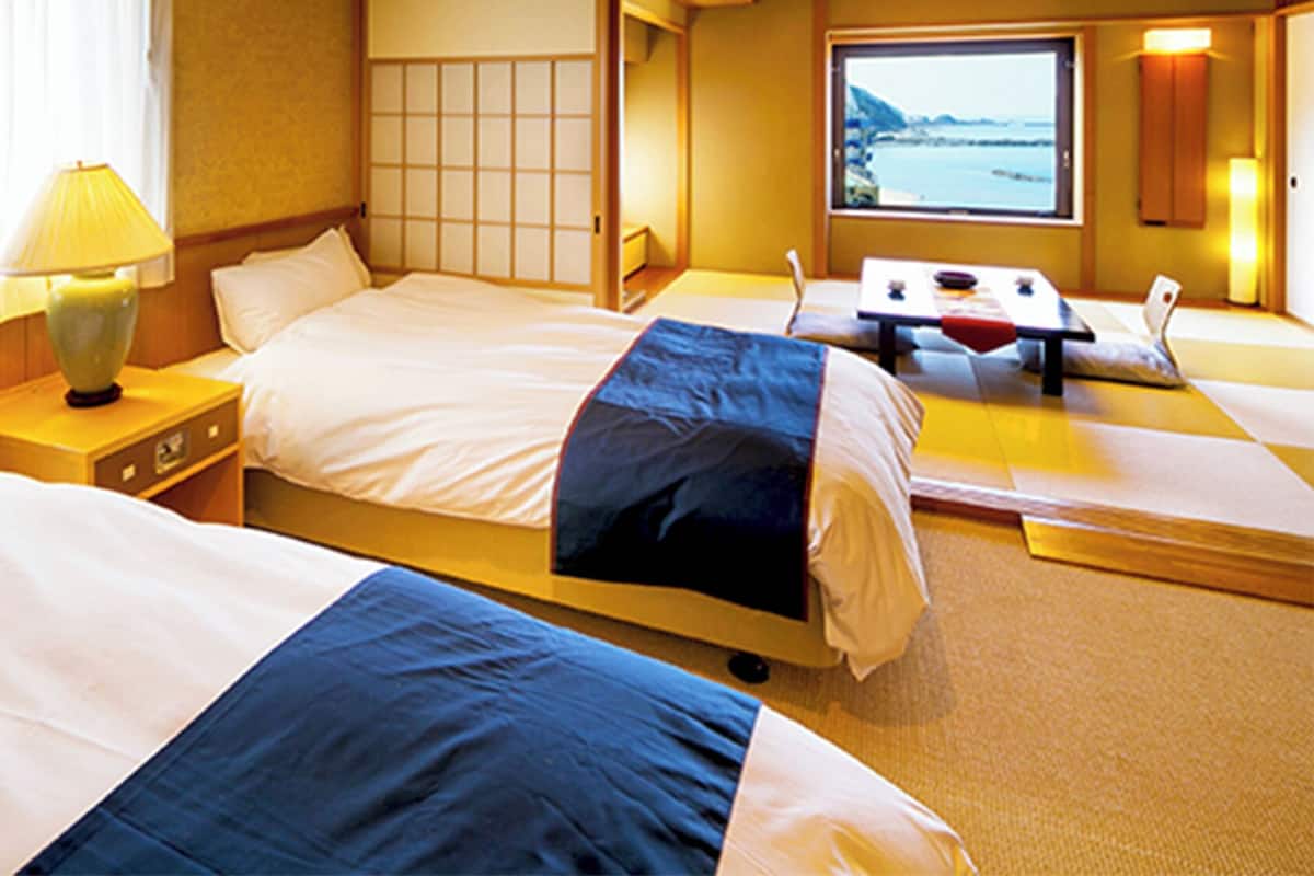 A hotel room with two beds, a bedside table with a lamp, a window with a view, and a small table with a chair.