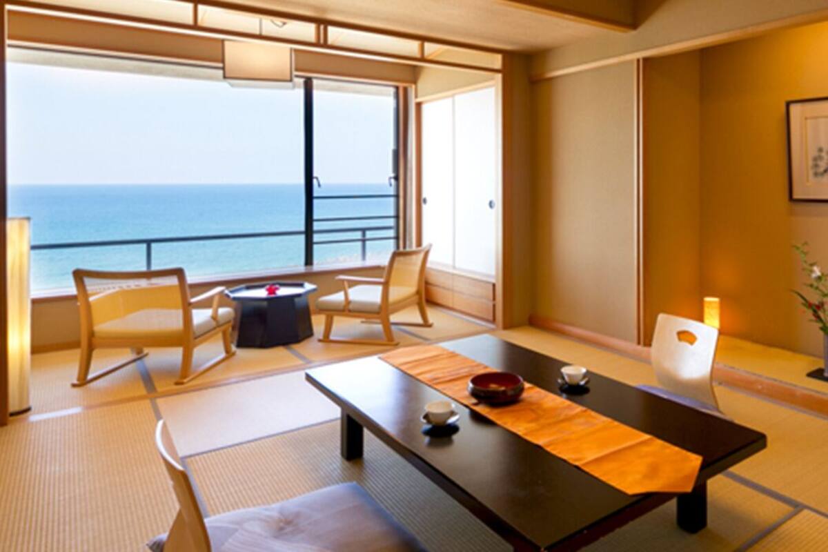 A traditional Japanese-style room with tatami flooring, a low table with a bowl and chopsticks, and a view of the sea through large sliding doors.