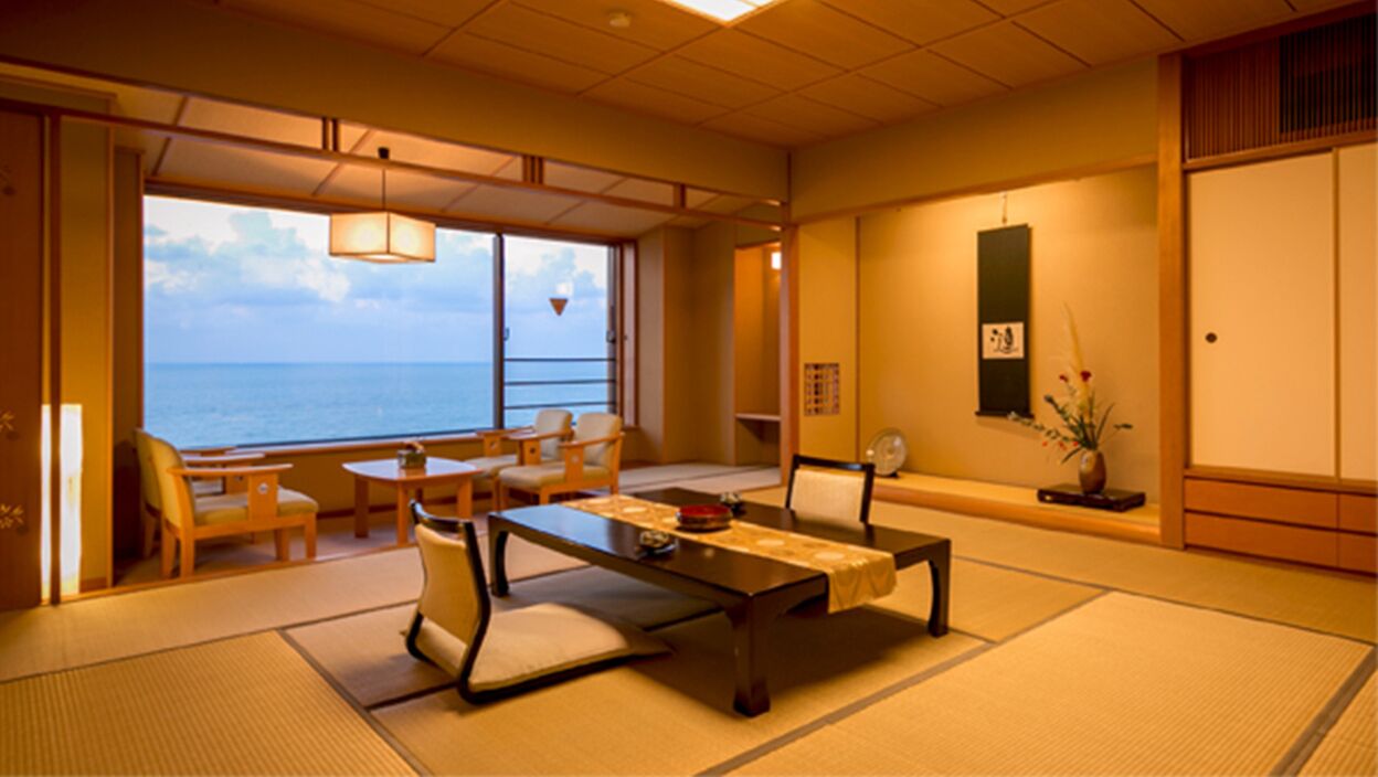A traditional Japanese room with tatami flooring, a low table with cushions, a sofa, and a view of the sea.