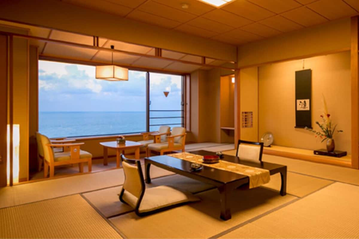 A traditional Japanese room with tatami flooring, a low table with cushions, a sofa, and a view of the sea.