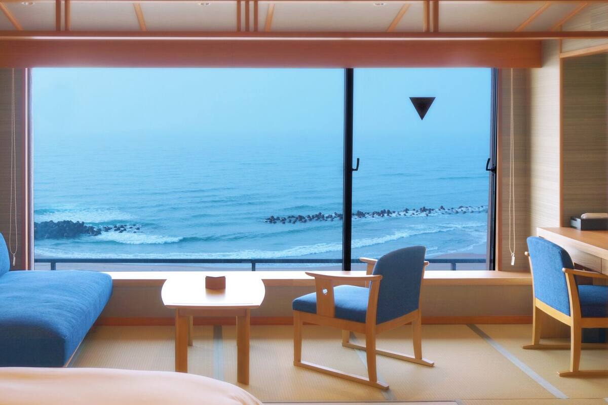 A room with a large window overlooking the sea, a sofa, a table, and a chair.