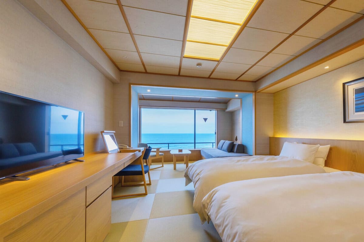 A hotel room with a bed, a desk with a TV, a window with a view of the sea, and a framed picture on the wall.