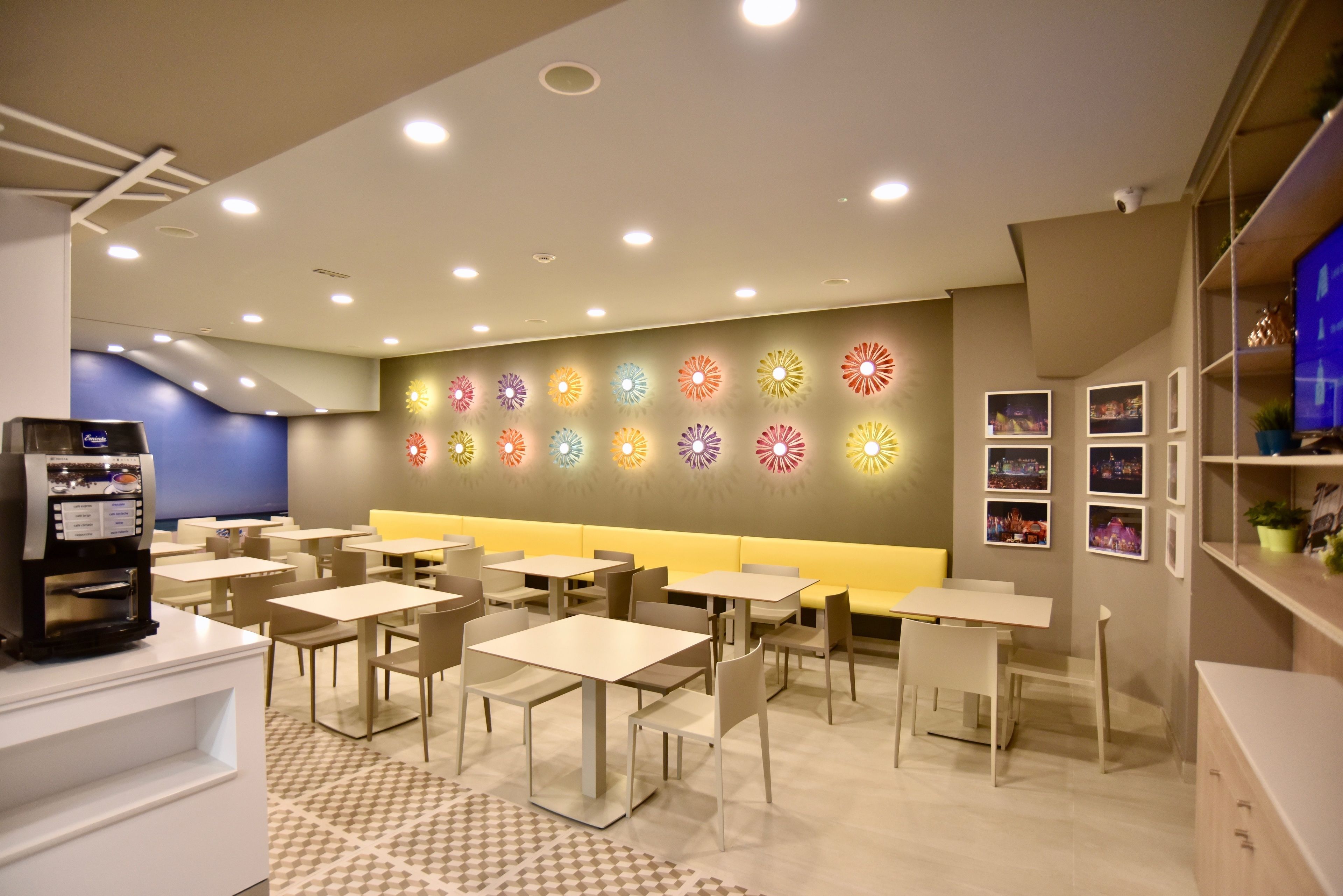 A modern dining area with multiple tables and chairs, a coffee machine, and a wall with colorful light fixtures.