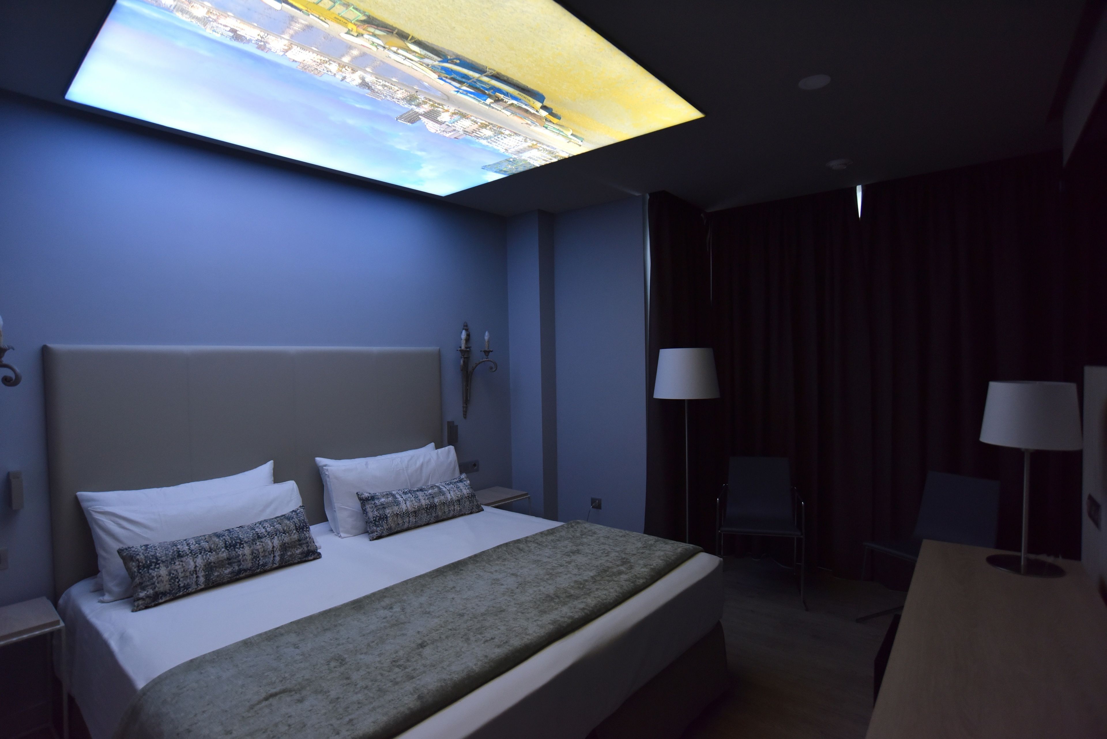 A hotel room with a bed, a desk, a chair, and a large window displaying a cityscape.