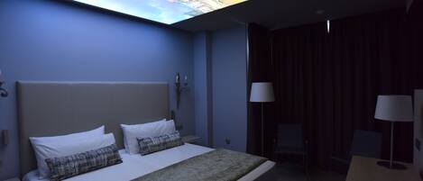 A hotel room with a bed, a desk, a chair, and a large window displaying a cityscape.