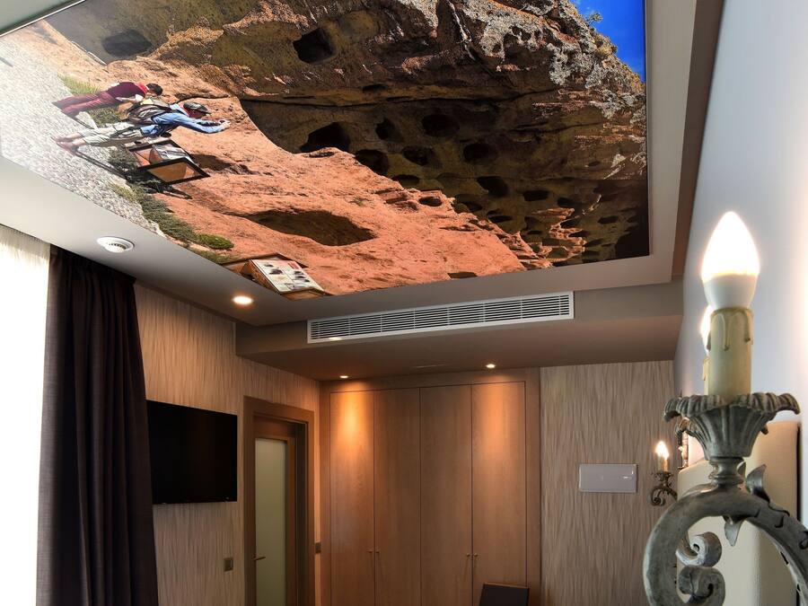 A hotel room with a bed, a television displaying a cave scene, a wooden wardrobe, a chair, and a wall-mounted light fixture.