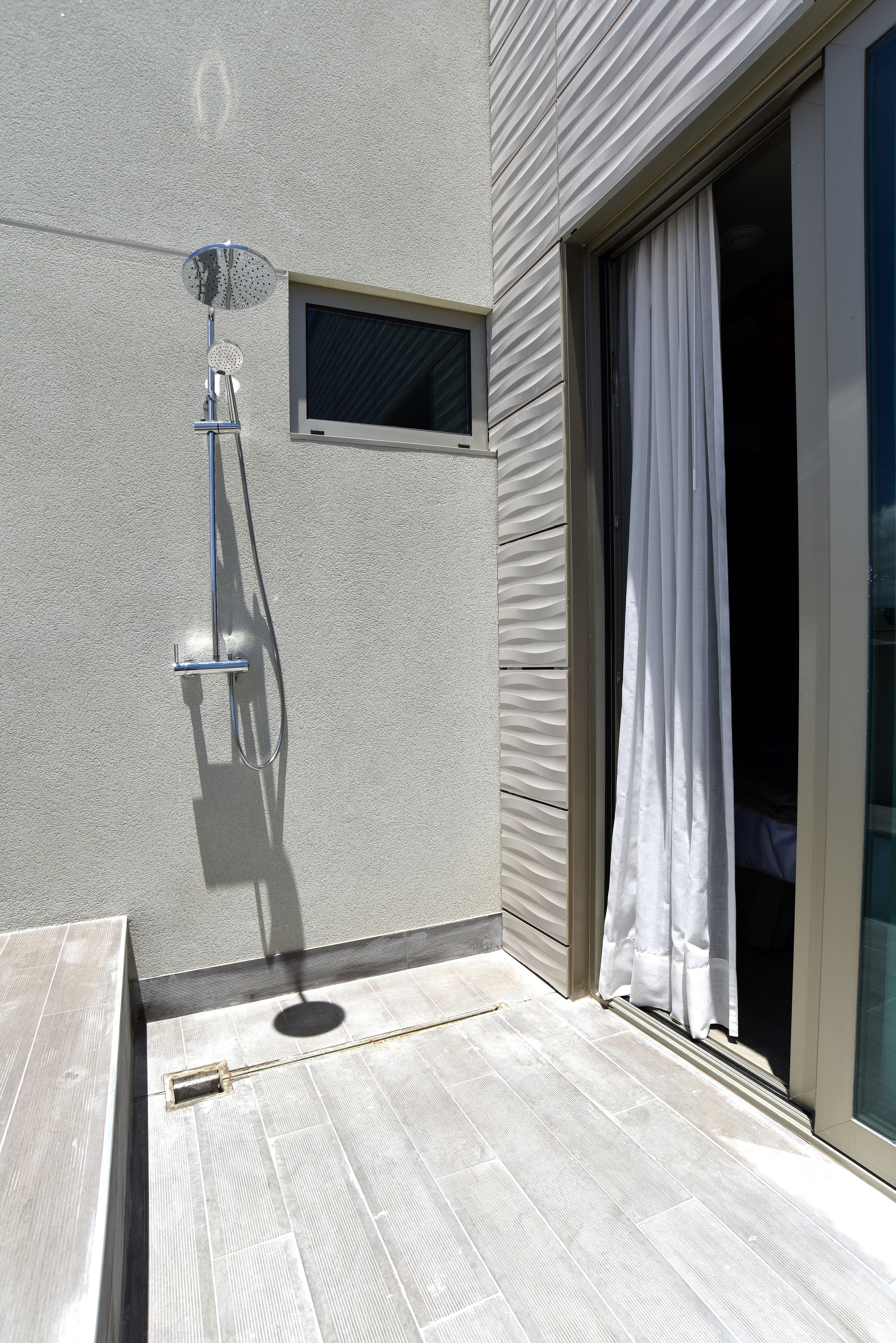 A modern outdoor shower area with a metal hose and showerhead, a small window, and a sliding glass door with a white curtain.