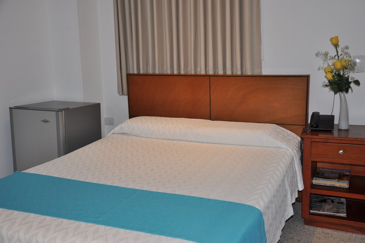 A neatly made bed with a blue accent, a wooden nightstand with a vase of flowers, and a small desk lamp.