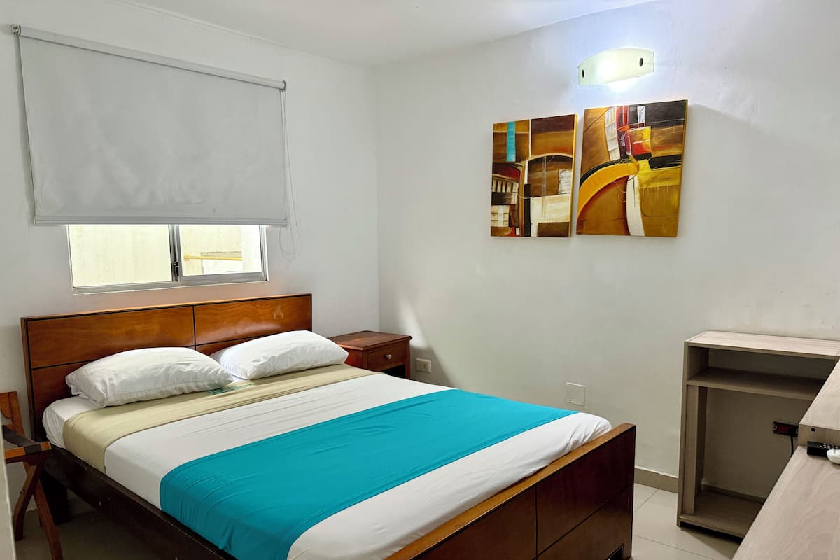 A neatly arranged bedroom with a wooden bed, a bedside table, a nightstand, a window with a blind, and a wall-mounted artwork.