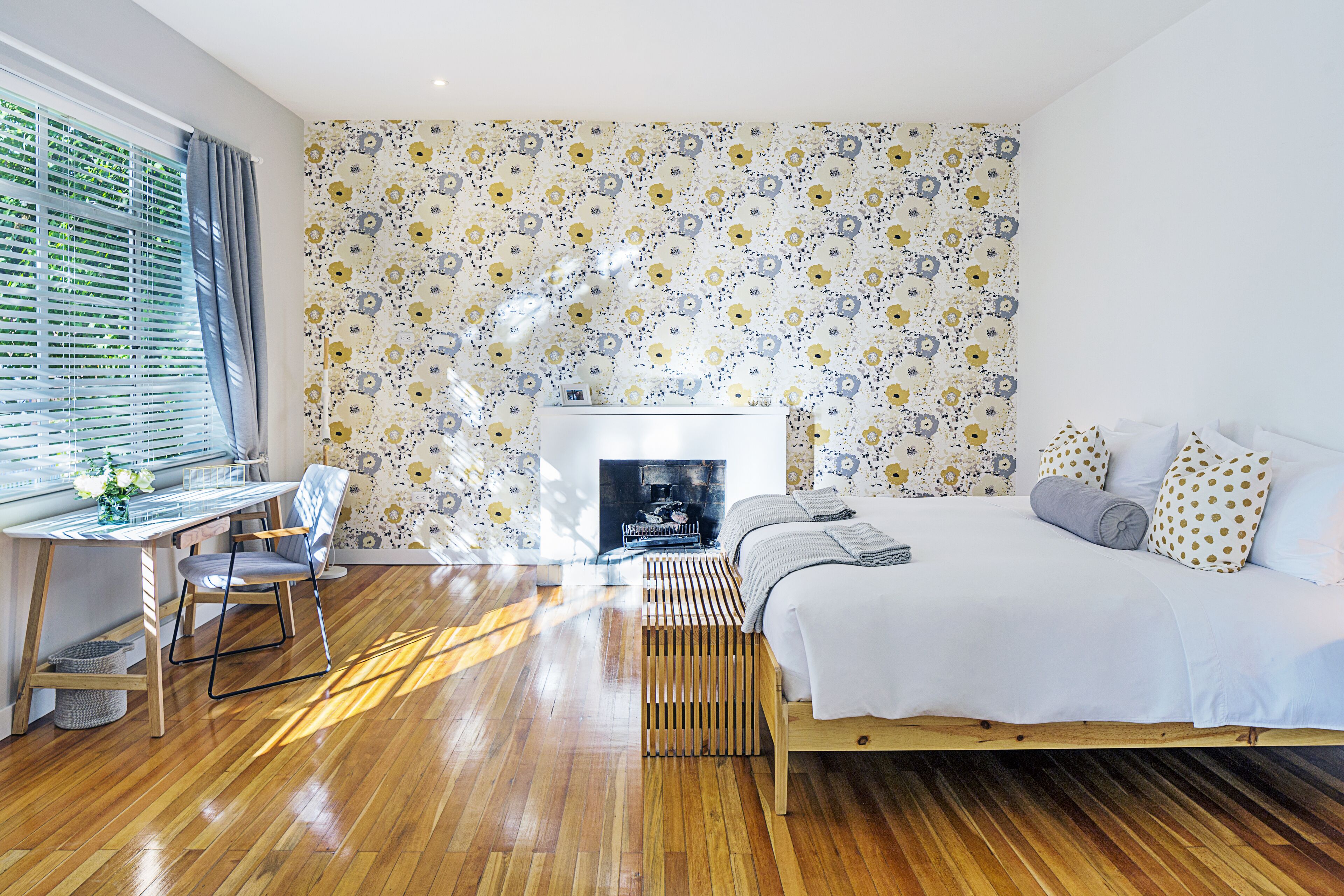 Une chambre avec un plancher en bois, un lit avec une literie blanche, un bureau avec une chaise et un papier peint à motifs.