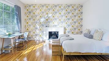 Une chambre avec un plancher en bois, un lit avec une literie blanche, un bureau avec une chaise et un papier peint à motifs.