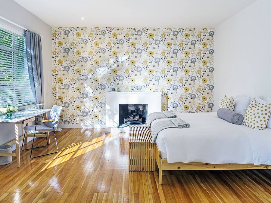 Une chambre avec un plancher en bois, un lit avec une literie blanche, un bureau avec une chaise et un papier peint à motifs.