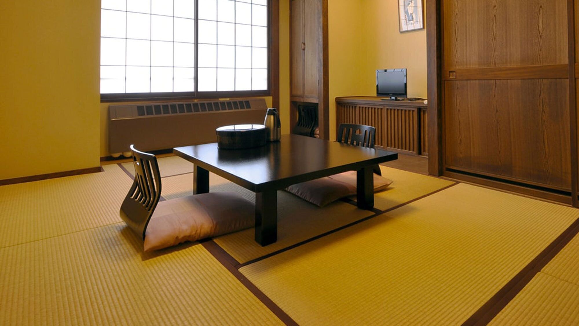 A traditional Japanese room with tatami flooring, a low table, cushions, a chair, and a television.