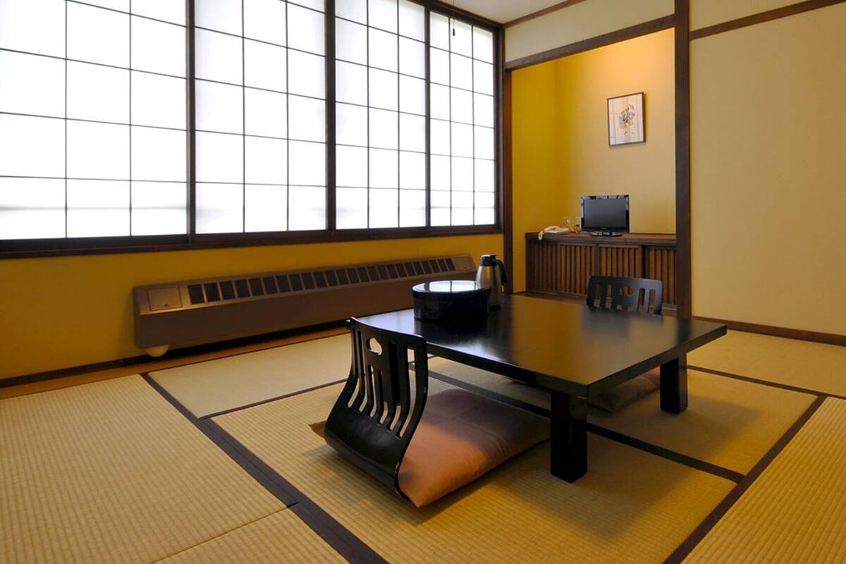 Una habitación japonesa tradicional con suelo de tatami, una mesa baja con cojines, un calentador y un televisor sobre un soporte.