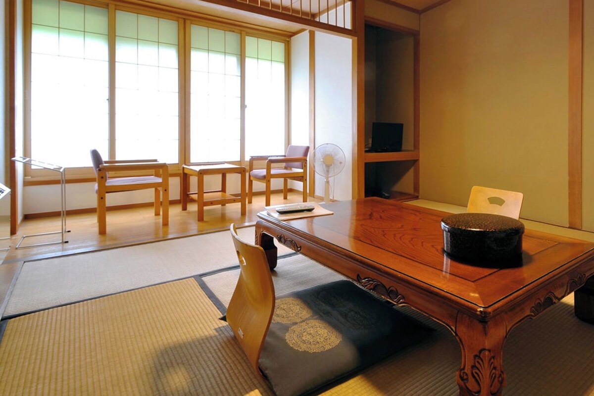 A tatami mat room with a low table, chairs, a fan, and a television.
