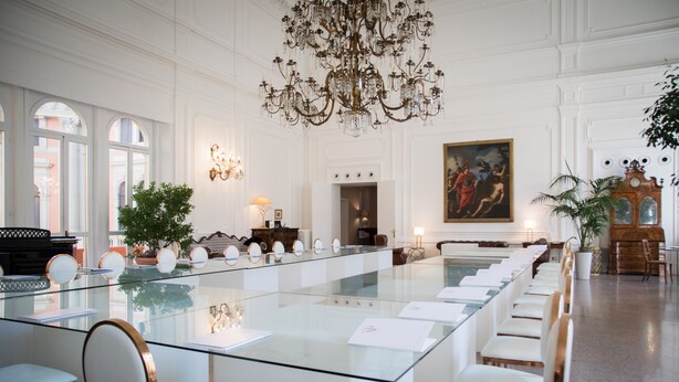 A spacious conference room with a long glass table, white chairs, and a chandelier.