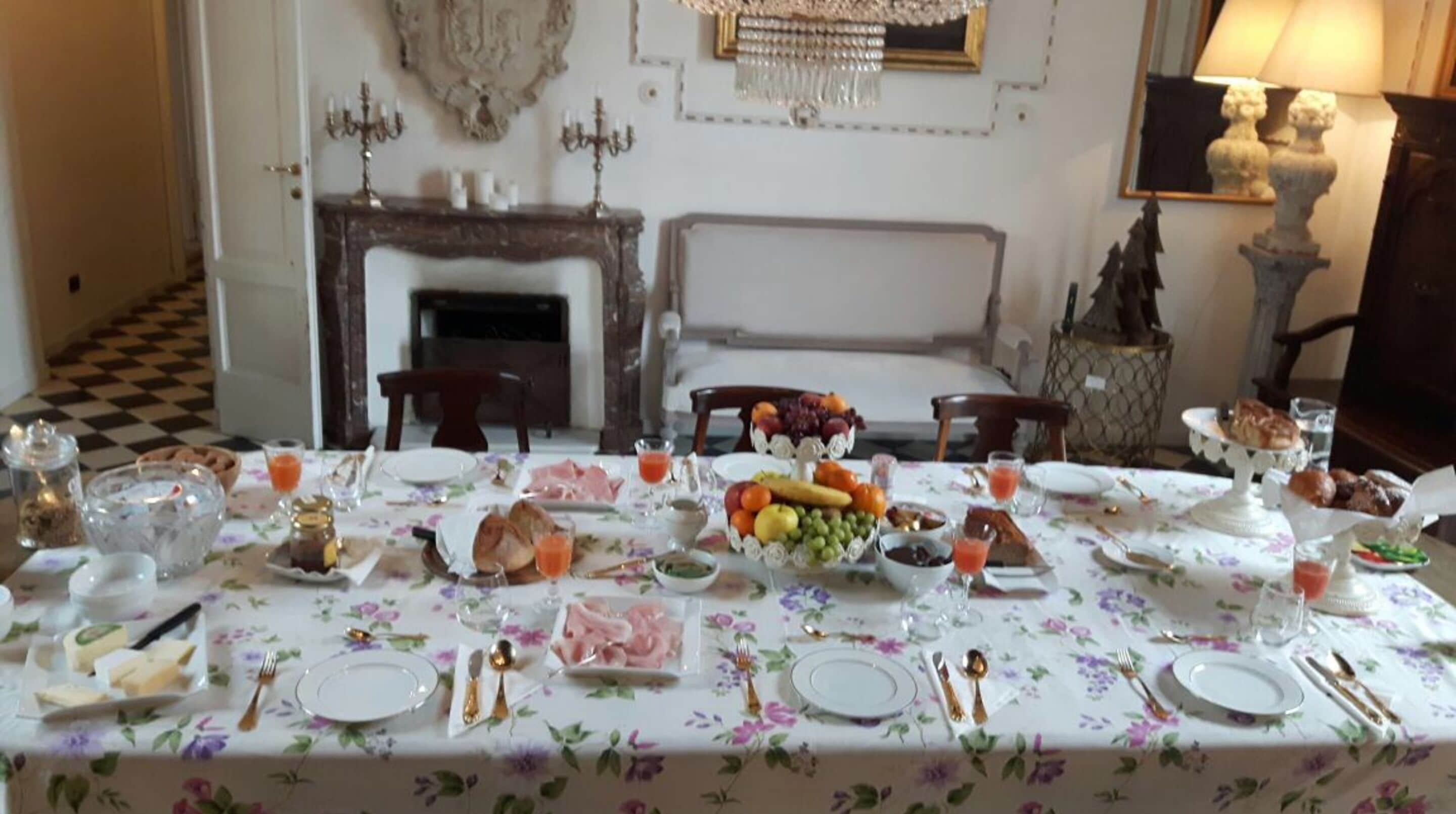 A dining table set with plates, cutlery, and a variety of food items, including fruits and bread, in a room with a fireplace and a chandelier.
