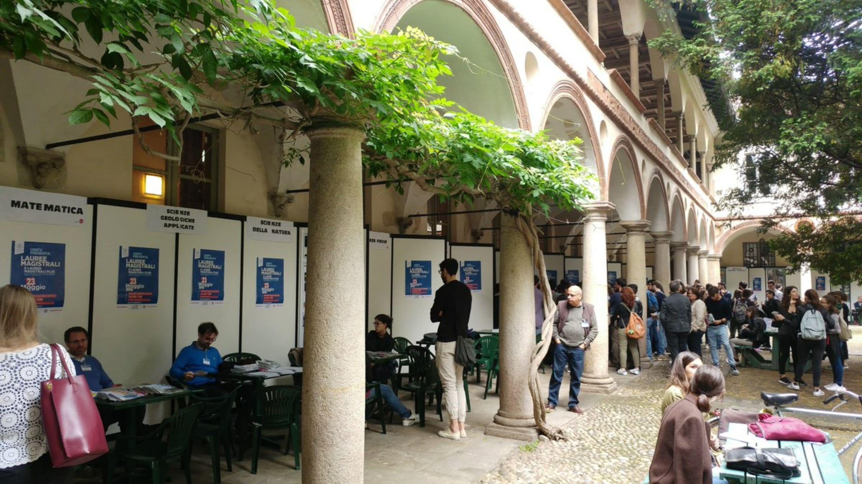Un área de estar al aire libre con mesas y sillas, rodeada de columnas y arcos, con gente sentada y de pie.
